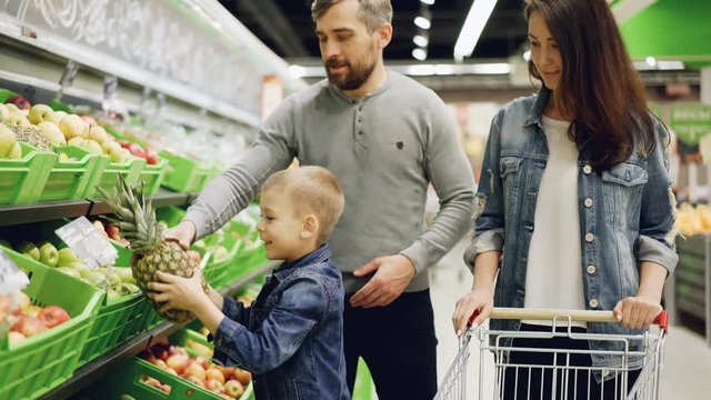 Happy young family mother, father and child are buying fruit in supermarket putting pineapple in shopping cart, talking and smiling. Food store and people concept.