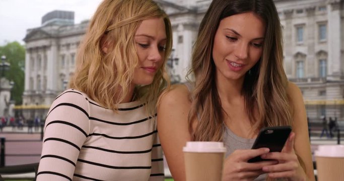 Close up of pretty brunette woman showing blonde friend smart phone in front Buckingham Palace in London, England, Close view of two friends sharing smartphone in London, 4k
