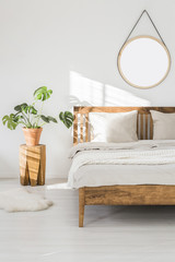 Monstera plant on a tree trunk night stand and a round mirror on a white wall in a sunlit bedroom interior with wooden furniture © Photographee.eu