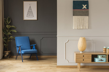 Blue armchair against grey wall with poster and wooden cabinet in living room interior. Real photo