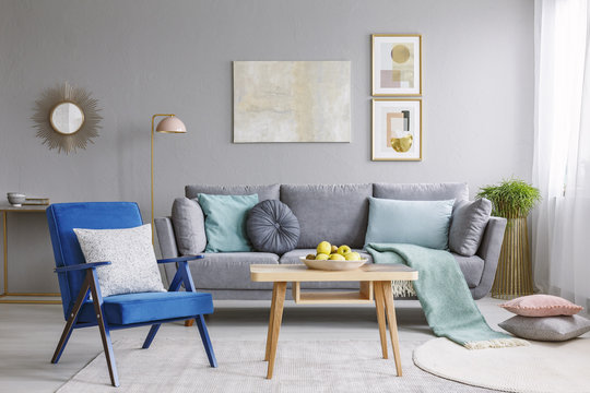 Blue armchair with pillows standing next to wooden table with fresh apples in grey living room interior with decorative mirror and posters hanging on the wall and grey lounge with cushions