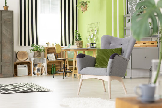 Green pillow on grey armchair in modern living room interior with striped drapes. Real photo