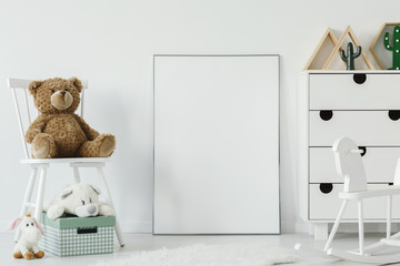 Teddy bear on white chair next to white poster with mockup in child's room interior. Real photo....