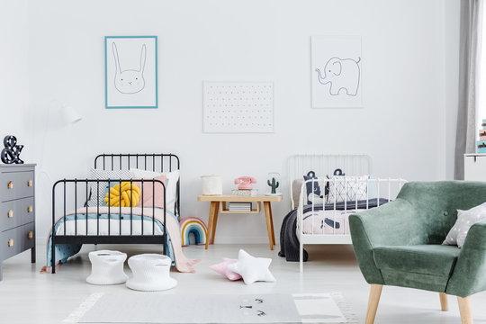 Bright scandinavian style bedroom interior with two metal frame kid's beds, one white, one black, and green armchair for caretaker. Posters of a rabbit and an elephant on white wall. Real photo