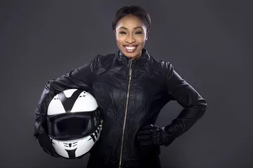 Fotobehang Zwarte vrouwelijke motorrijder of autocoureur of stuntvrouw die een leren racepak draagt en een beschermende helm vasthoudt. Ze staat zelfverzekerd in een studio © Innovated Captures