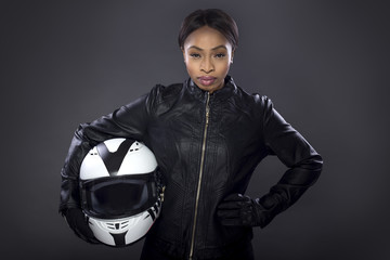 Black female motorcycle biker or race car driver or stuntwoman wearing leather racing suit and holding a protective helmet.  She is standing confidently in a studio