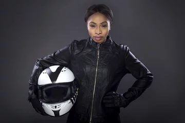 Rolgordijnen Black female motorcycle biker or race car driver or stuntwoman wearing leather racing suit and holding a protective helmet.  She is standing confidently in a studio © Innovated Captures