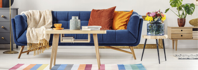 Closeup of a wooden table and a blue settee with orange pillows standing next to a bouquet of spring flowers on a stool in bright living room interior. Real photo