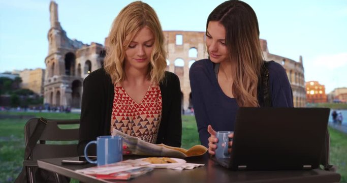 Cute blonde girl showing friend map of Europe while sitting in front Colosseum with laptop computer in Rome, Italy, Two beautiful young woman sitting with travel map near ancient Roman landmark, 4k