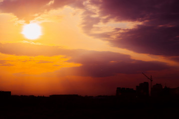 Magenta sunrise above silhouette of city buildings with tower cranes. Unusual cityscape with beautiful dawn. Picturesque warm city landscape with bright sunny sky. Big sun above city architecture.