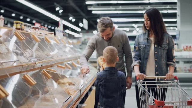 Adorable little boy is helping his parents to choose bread in bakery department in food store, he is taking loaf and smelling it then putting in cart.