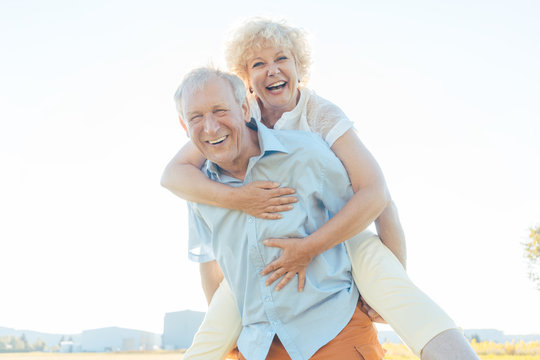 Low-angle view portrait of a happy senior man laughing, while carrying his partner on his back in a sunny day of summer in the countryside
