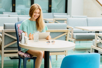 Communication, technology, leisure and people concept - positive young blond woman or teenage girl using tablet pc computer at modern restaurant or cafe terrace.
