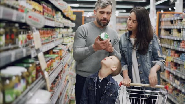 Married couple with cute little son are shopping for food, they are taking glass can from shelf and checking ingredients and expiry date and talking.