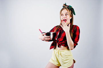 Young funny housewife in checkered shirt and yellow shorts pin up style with saucepan and kitchen spoon isolated on white background.