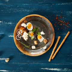 Bruschetta with cream cheese, fried eggs, mushrooms and salad leaves lying on plate standing on blue aged wooden surface with quail eggs and bread sticks scattered around. Tasty daily meal. Top view.