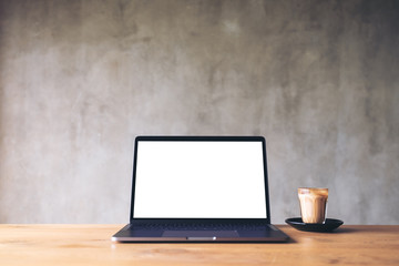 Mockup image of laptop with blank white desktop screen and coffee cup on wooden table with concrete wall background