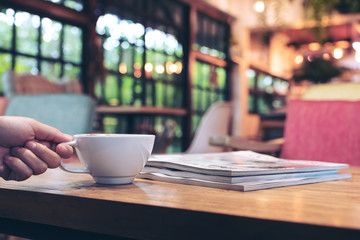 Closeup image of a hand holding white cup of hot coffee with the books on wooden table in cafe