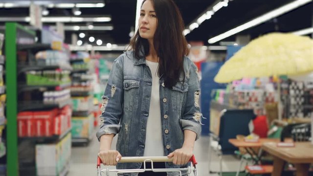 Attractive woman is driving shopping trolley through food department in supermarket and looking around. Pretty girls, everyday life and buying products concept.