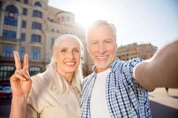 Hi! Rest relax leisure concept. Self portrait of friendly peaceful grandma and granddad shooting selfie on front camera over blurred buildings having video-call gesturing v-sign with two fingers