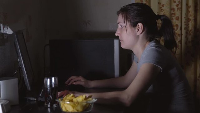 Young woman sitting at the computer and eating chips.
