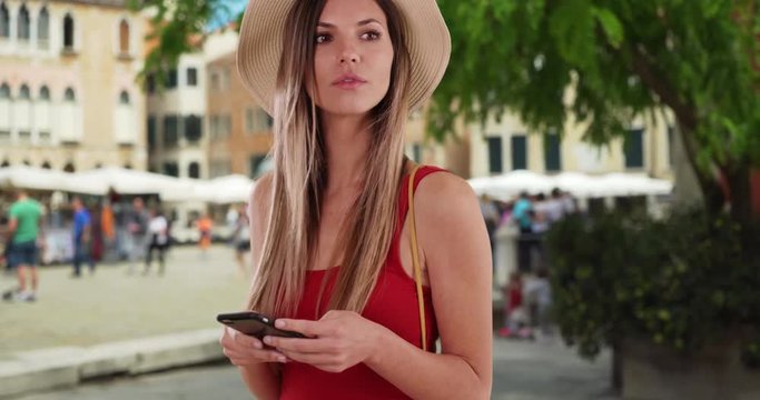 Stylish woman in red top using phone and looking around while in Venice, Italy, Caucasian hipster girl in her 20s texting on smartphone in Italian city setting, 4k