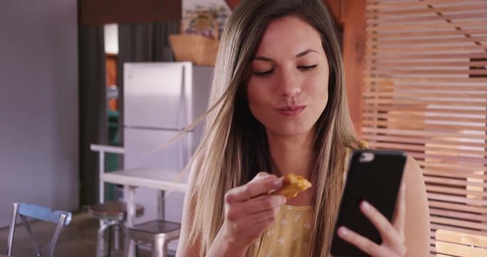 Close-up of girl taking selfie with phone while eating pizza in modern kitchen, Pretty woman taking selfie photo and eating slice of pizza in domestic kitchen setting, 4k