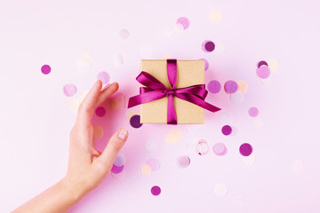 Woman hand and kraft gift box with pink bow on pink background decorated with confetti.. Top view,...