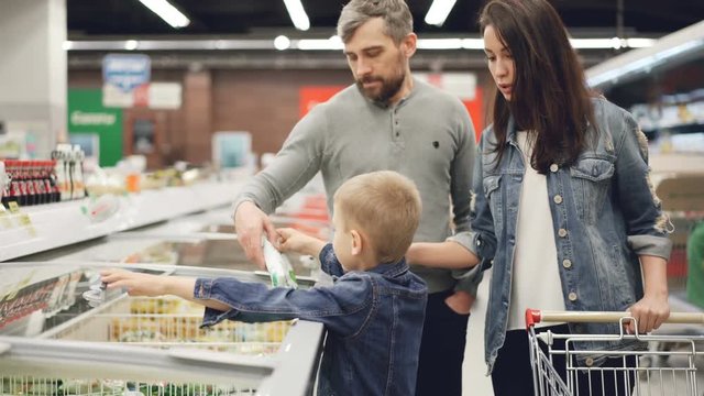 Little Boy Is Opening Freezer And Taking Pack Of Frozen Vegetables Then Giving It To His Daddy While Shopping In Supermarket With His Family. People And Shop Concept.