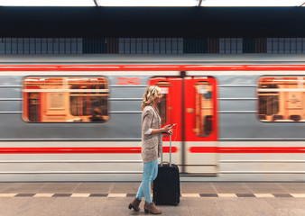 Young woman with suitcase looking at phone while waiting train in metro station.