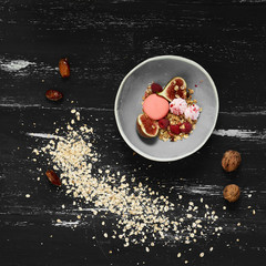 Pink macaron, meringues, fig, raspberries, cereals in bowl standing on blue aged wooden table with ground oats and whole walnuts scattered around. Tasty morning meal, delicious sweet food. Top view.