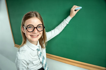 Portrait of cute schoolgirl in round glasses writing on blackboard in classroom. Education concept