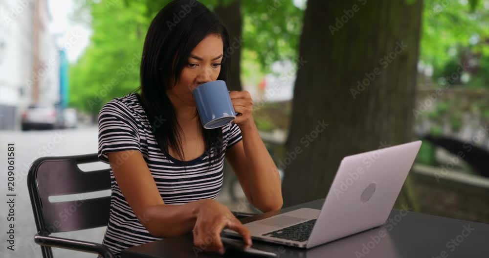 Wall mural Black woman working on laptop outside and drinking coffee, African American female with coffee writing email to her boss, 4k