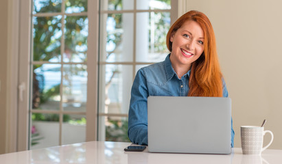 Redhead woman using computer laptop at home with a happy face standing and smiling with a confident smile showing teeth