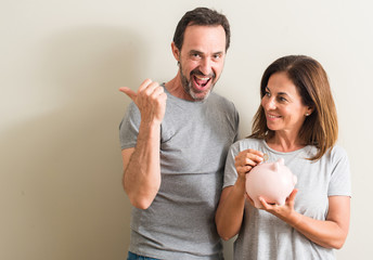 Middle age couple, woman and man, holding piggy bank pointing with hand and finger up with happy face smiling