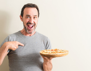 Senior man holding pizza with surprise face pointing finger to himself