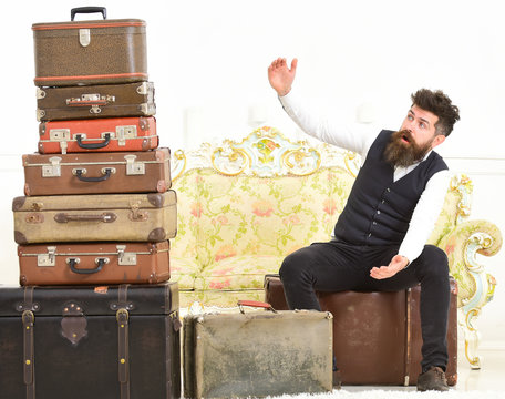Man, butler with beard and mustache delivers luggage, luxury white interior background. Luggage and travelling concept. Macho elegant on surprised face sits shocked near pile of vintage suitcase.