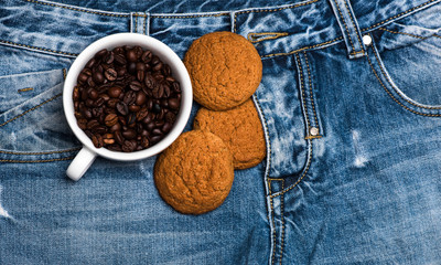 Cup with coffee beans and homemade oat cookies lay on zip of jeans, denim background. Coffee break concept. Mug full of coffee beans and oat cookie on jeans