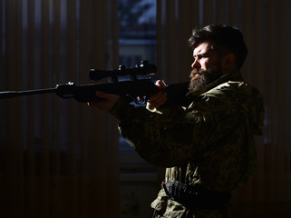 Macho on suffering grimace face aiming at victim. Hunter, soldier with gun aiming before shooting. Shooter concept. Man with beard wears camouflage clothing, dark interior background.