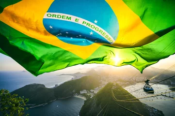 Poster de jardin Brésil Le drapeau brésilien brille au-dessus des toits de la ville au coucher du soleil doré à Sugarloaf Mountain à Rio de Janeiro au Brésil.