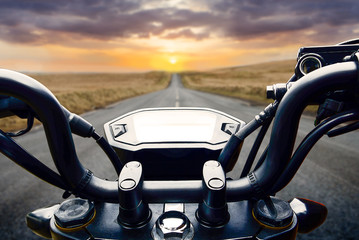 Motorcycle handlebars riding toward sunset horizon on lonely straight road.