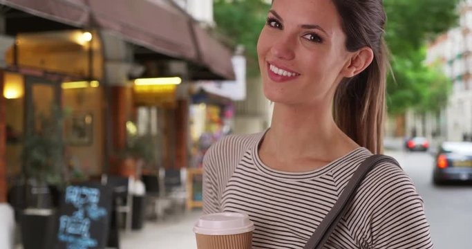 Portrait of smiling millennial woman with coffee out on sidewalk by cafe, Young Caucasian lady wearing striped shirt smiling at camera outside by coffee shop, 4k