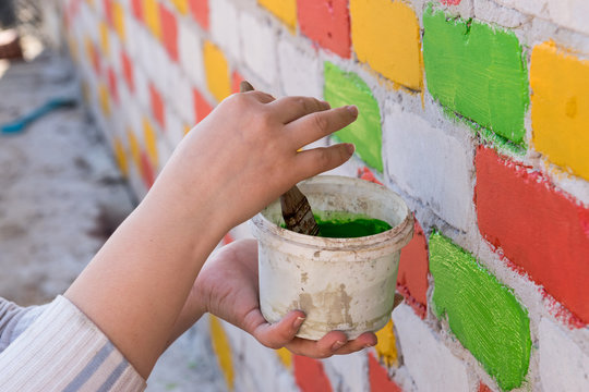 Young Caucasian shapely woman paints a brick wall with paint brush in different colors. Red, blue, yellow, green. Capacity with paint and hands close-up view
