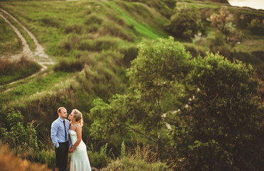 Young bride and groom with curly hair embrace against the background of green rocks and hills with backlight. Evening wedding of the newlyweds. Newlyweds at sunset.
