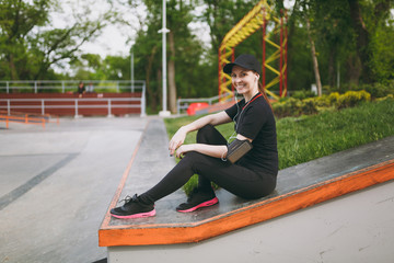 Young athletic smiling beautiful girl in black uniform and cap with headphones listening music, resting and sitting before or after running, training in city park outdoors. Fitness, healthy lifestyle.