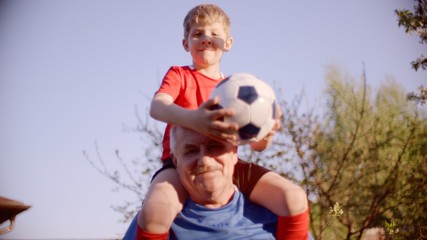 Little boy is sitting on the neck of his grandfather and holding soccer ball in his hands