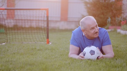 Old man wearing football uniform is lying on the lawn with soccer ball