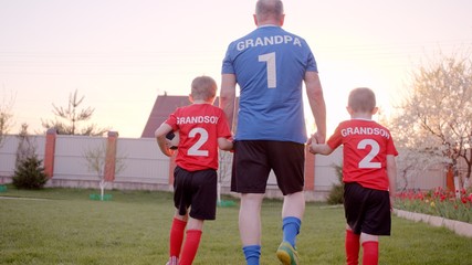 Backshot of grandfather is handling his grandsons, wearing football uniform, with soccerball