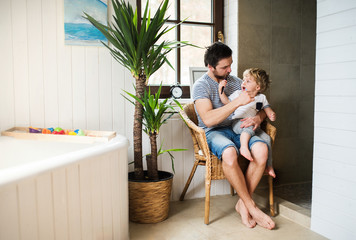 Father and a toddler boy sitting on a chair and brushing their teeth at home.