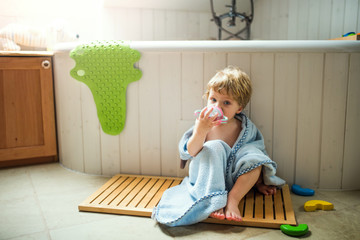 A toddler boy wrapped in towel sitting on the floor in the bathroom at home.